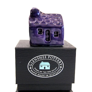 May include: A small purple ceramic house with a thatched roof, sitting on a black box with a white label that reads "Glenshee Pottery, Home of the Wee Bothy, Hand Made in Scotland".