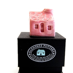 May include: A small pink ceramic house with a white chimney and a black box with the text "Glenshee Pottery, Home of the Wee Bothy, Hand Made in Scotland".