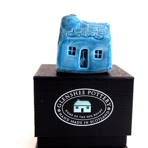 May include: A small blue ceramic house with a white door and windows, sitting on a black box with a white label that reads "Glenshee Pottery, Home of the Wee Bothy, Hand Made in Scotland".