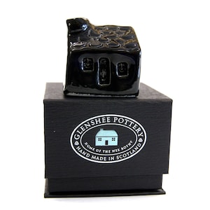 May include: A glossy black ceramic miniature house figurine with a textured roof and three windows, sitting atop a black box. The box features a white and blue logo that reads "Glenshee Pottery, Home of the Wee Bothy, Hand Made in Scotland."