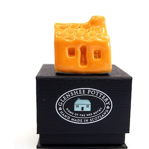 May include: A small orange ceramic house with a thatched roof, sitting on a black box with the text "Glenshee Pottery, Home of the Wee Bothy, Hand Made in Scotland".