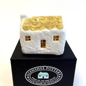 May include: A small white ceramic cottage with a yellow roof, sitting on a black box with the text "Glenshee Pottery Home of the Wee Bothy Hand Made in Scotland".