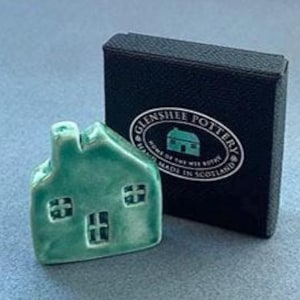 May include: A small, turquoise ceramic house with a chimney and windows, next to a black box. The box has a white circular logo with the text "Glenshee Pottery" and "Home of the Wee Bothy, Made in Scotland."
