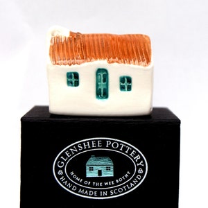 May include: A small white ceramic cottage with a brown roof and teal windows. The cottage is sitting on a black box with a white label that reads "Glenshee Pottery, Home of the Wee Bothy, Hand Made in Scotland".