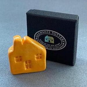 May include: A small, orange ceramic house figurine with a pitched roof and window details. Beside it is a black box with the "Glenshee Pottery" logo. The figurine is likely a decorative item or collectible.