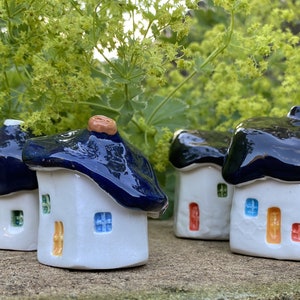 Puede incluir: Cuatro pequeñas casas de cerámica con techos azules y detalles coloridos. Las casas son blancas con ventanas y puertas de diferentes colores. Están dispuestas en fila sobre una superficie marrón.