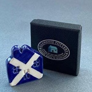 May include: A small, ceramic house figurine with a blue and white Scottish flag design. The figurine is next to a black box with the text "Glenshee Pottery, Home of the Wee Bothy, Hand Made in Scotland."