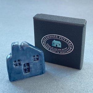 May include: A small, blue ceramic house figurine with three windows and a chimney, next to a black box with the text "Glenshee Pottery, Home of the Wee Bothy, Hand Made in Scotland" printed on it. The house is approximately 2 inches tall.