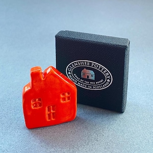May include: A small, vibrant orange ceramic house with embossed windows and a chimney. The house is next to a black box with the text "Glenshee Pottery, Home of the Wee Bothy, Hand Made in Scotland" printed on it.