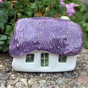 Könnte beinhalten: Ein kleines, weißes Keramikhaus mit einem violetten Strohdach und grünen Fenstern. Das Haus steht auf einem Bett aus kleinen Steinen.