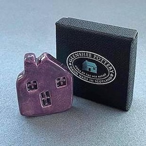 May include: A small, glossy, purple ceramic house figurine with three windows and a chimney, next to a black box with a white label that reads "Glenshee Pottery, Hand Made in Scotland."