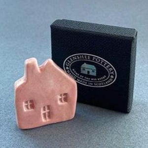 May include: A small, pink ceramic house with three windows and a chimney, next to a black box with the text "Glenshee Pottery" and "Handmade in Scotland". The house is a decorative item.