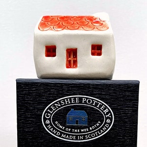 May include: A small white ceramic house with a red roof and three windows. The house is sitting on a black box with the text "Glenshee Pottery Home of the Wee Bothy Hand Made in Scotland" printed on it.