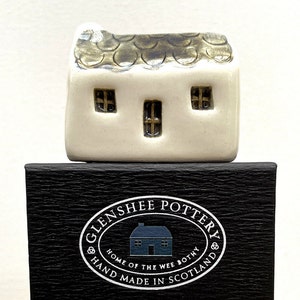 May include: A small white ceramic house with a brown roof and three windows. The house is sitting on a black box with the words "Glenshee Pottery" and "Home of the Wee Bothy" printed on it.
