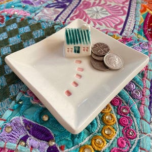 May include: A square, cream-coloured ceramic trinket dish featuring a miniature house and coins. The house has a green roof. A series of pink dots lead to the house. The dish is on a colourful, patterned textile. Coins are visible on the dish.