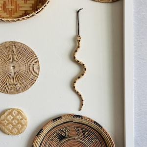 May include: A light-colored wooden snake ornament featuring dark brown geometric patterns along its body. The snake has a curved, S-shaped posture and is suspended from a thin black cord attached to its head for hanging purposes.