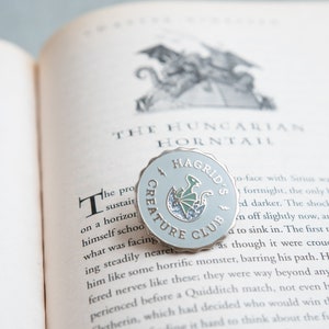 May include: A light blue Hagrid's Creature Club pin with a scalloped edge, featuring a green dragon hatching from an egg. The pin is set on an open book with the text "THE HUNGARIAN HORNTAIL".