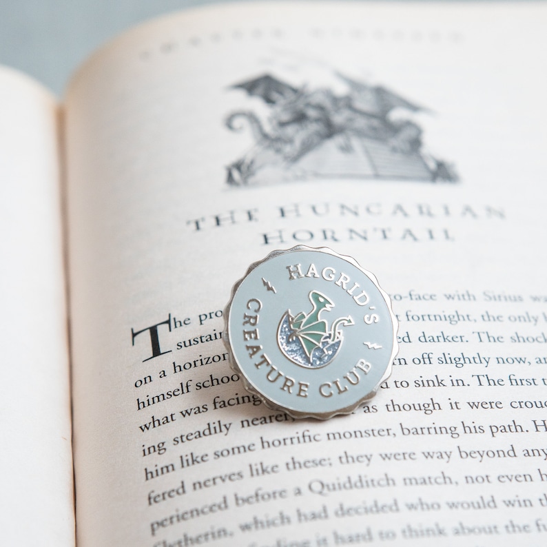May include: A light blue Hagrid's Creature Club pin with a scalloped edge, featuring a green dragon hatching from an egg. The pin is set on an open book with the text "THE HUNGARIAN HORNTAIL".