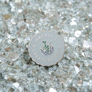 May include: A round, light gray enamel pin with the text "HAGRID'S CREATURE CLUB" and a green and black dragon design. The pin is set against a background of clear, crushed glass.