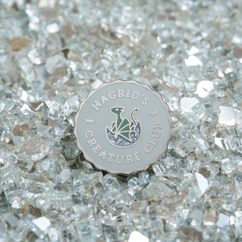 May include: A round, light gray enamel pin with the text "HAGRID'S CREATURE CLUB" and a green and black dragon design. The pin is set against a background of clear, crushed glass.