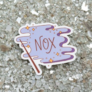 May include: A sticker with a light purple cloud design, the word "NOX" in brown script, and a brown wand. The design includes gold stars and purple accents. The sticker is on a reflective, textured surface.