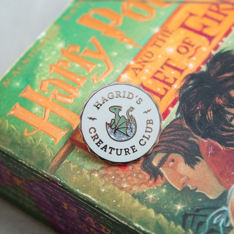 May include: A round enamel pin with the text "Hagrid's Creature Club" and a green dragon hatching from an egg. The pin has a white background with a silver glitter base and a gold-colored rim. The pin is placed on a book.