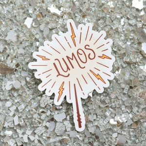 May include: A white sticker with the word "LUMOS" in brown script, surrounded by radiating lines and yellow lightning bolts. A brown wand extends from the bottom. The sticker is on a reflective, textured surface.