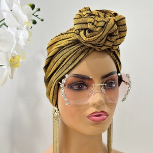 Pode incluir: Um turbante listrado em dourado e preto com um grande n&oacute; no topo. O turbante &eacute; usado por um manequim com &oacute;culos de sol transparentes adornados com strass e brincos de corrente dourada.