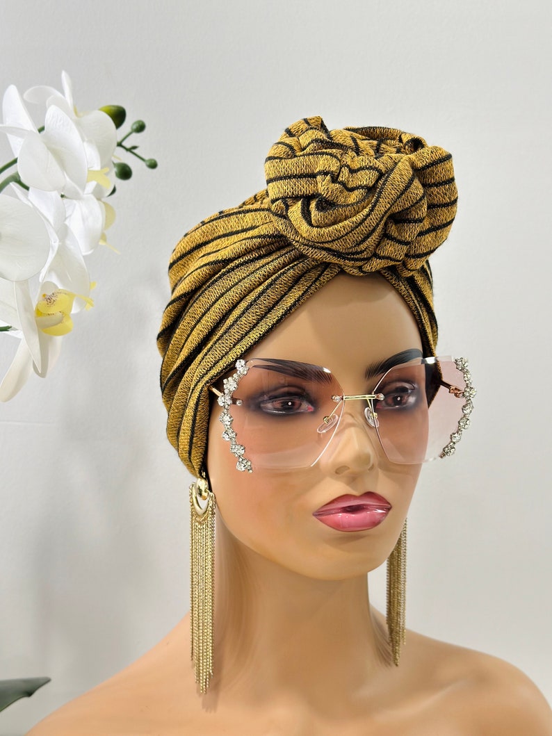 Pode incluir: Um turbante listrado em dourado e preto com um grande n&oacute; no topo. O turbante &eacute; usado por um manequim com &oacute;culos de sol transparentes adornados com strass e brincos de corrente dourada.