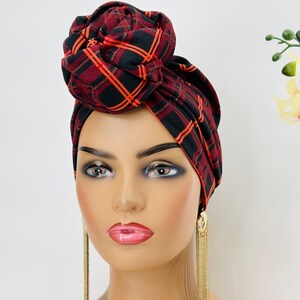 Pode incluir: Um turbante xadrez vermelho e preto com um grande n&oacute; no topo. O turbante &eacute; usado em um manequim com longos brincos dourados pendurados.