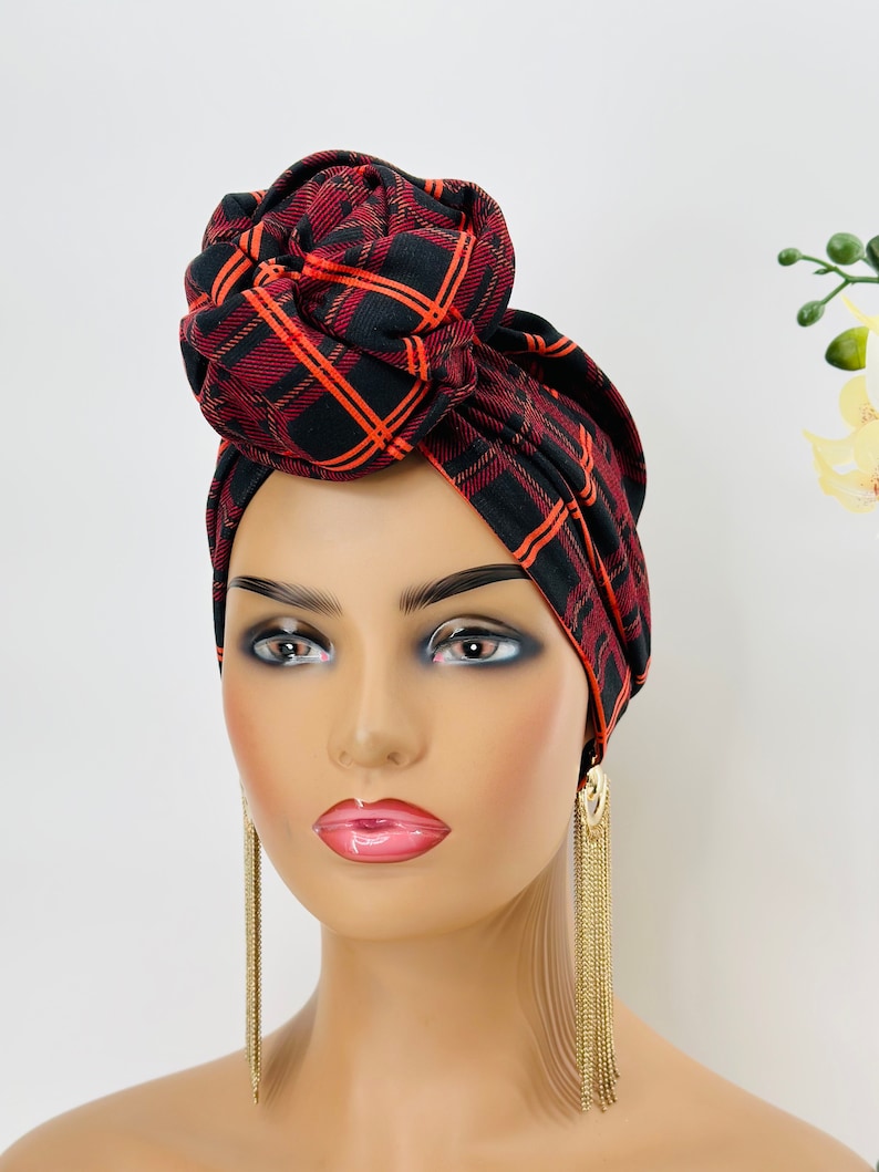Pode incluir: Um turbante xadrez vermelho e preto com um grande n&oacute; no topo. O turbante &eacute; usado em um manequim com longos brincos dourados pendurados.