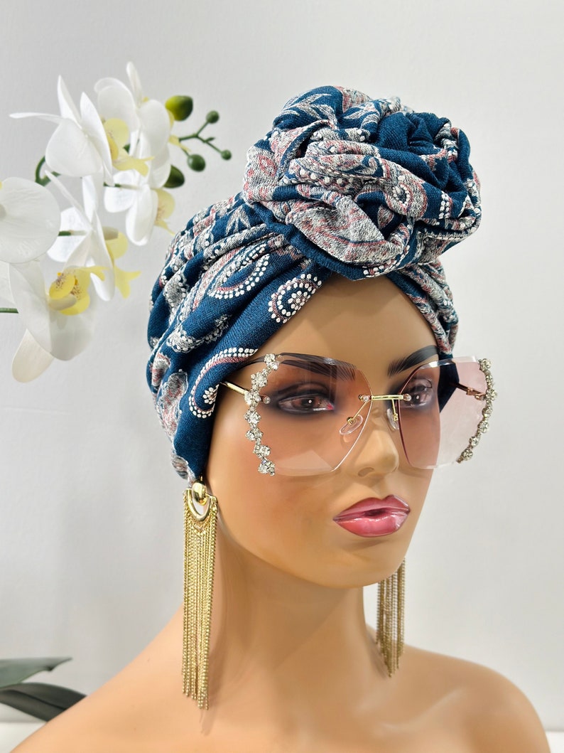 Pode incluir: Um turbante azul e branco estampado com um grande n&oacute; no topo. O turbante &eacute; usado por um manequim com longos brincos de corrente dourada e grandes &oacute;culos de sol transparentes adornados com strass.