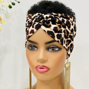 May include: A brown and white leopard print turban headband with a twist design. The headband is worn on a mannequin with short, curly black hair.