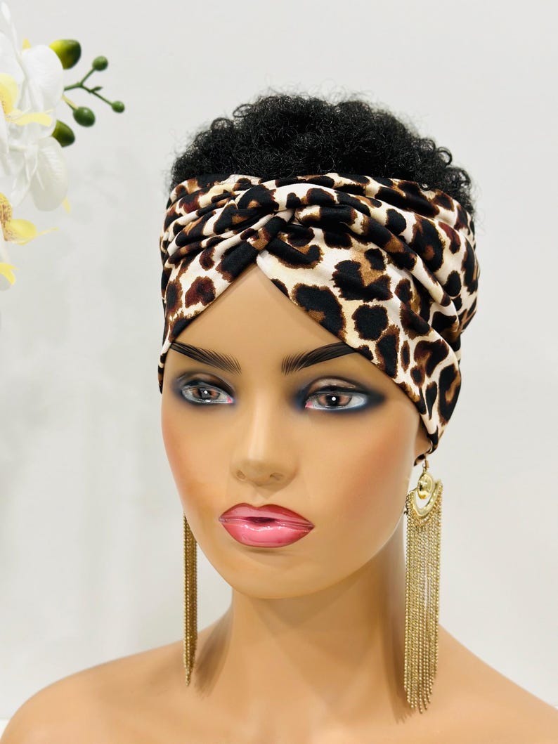 May include: A brown and white leopard print turban headband with a twist design. The headband is worn on a mannequin with short, curly black hair.