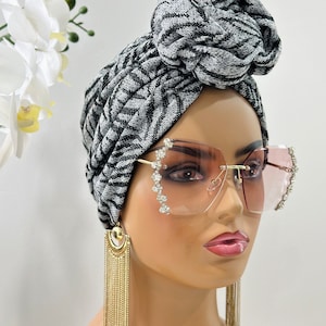 Pode incluir: Um turbante com padr&atilde;o preto e branco com um n&oacute; no topo. O turbante &eacute; usado por um manequim com &oacute;culos de sol rosa e brincos de corrente dourada.