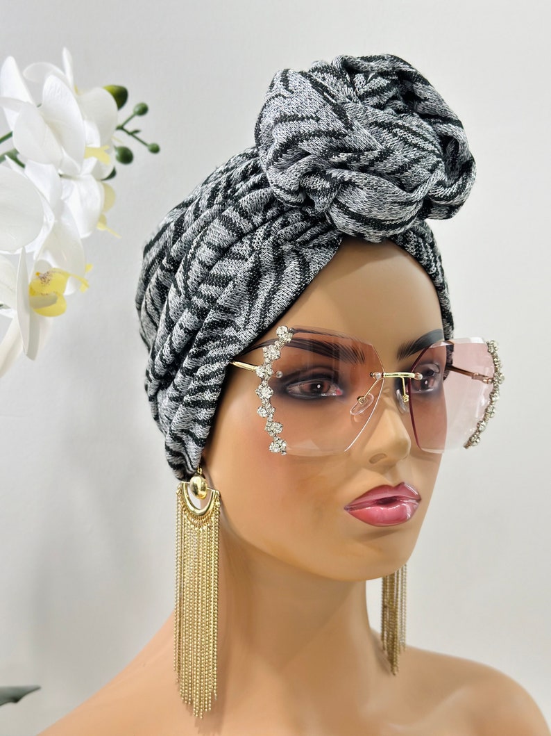 Pode incluir: Um turbante com padr&atilde;o preto e branco com um n&oacute; no topo. O turbante &eacute; usado por um manequim com &oacute;culos de sol rosa e brincos de corrente dourada.