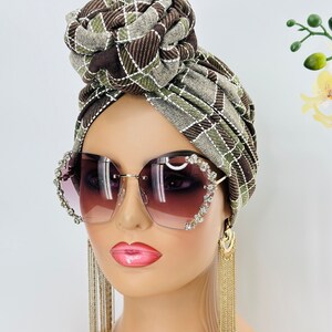 Pode incluir: Um turbante estampado em marrom e verde com um n&oacute; no topo. O turbante &eacute; usado por um manequim com cabelo castanho e que usa &oacute;culos de sol grandes em forma de cora&ccedil;&atilde;o com pedrarias e brincos de corrente dourada.