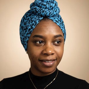 Può includere: Una donna indossa un turbante blu con un motivo animalier nero. Il turbante è avvolto a forma di chignon sulla sommità della testa. Indossa un dolcevita nero e una collana d'oro.