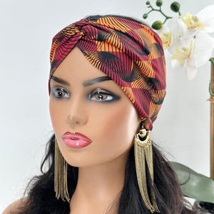 May include: A red and orange patterned turban headband with a knot on top. The headband is made of a soft, stretchy fabric.