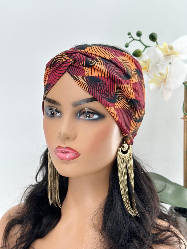 May include: A red and orange patterned turban headband with a knot on top. The headband is made of a soft, stretchy fabric.