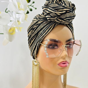 Pode incluir: Um turbante listrado preto e dourado com um n&oacute; no topo. O turbante &eacute; usado por uma pessoa que usa &oacute;culos de sol rosa com strass e brincos de corrente dourada.
