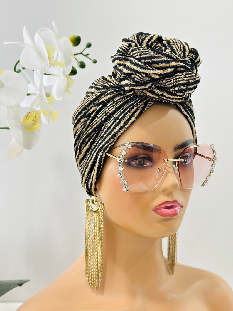 Pode incluir: Um turbante listrado preto e dourado com um n&oacute; no topo. O turbante &eacute; usado por uma pessoa que usa &oacute;culos de sol rosa com strass e brincos de corrente dourada.