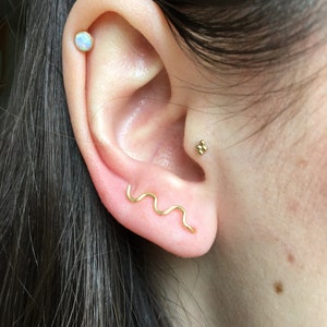 May include: A close-up of a person's ear with three gold earrings. The top earring is a small, round opal. The middle earring is a small cluster of gold balls. The bottom earring is a thin, wavy gold bar.