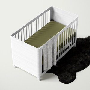 May include: A white wooden crib with a green fitted sheet and a white patterned blanket hanging over the side. The crib is sitting on a black fluffy rug.