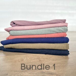 May include: A stack of six folded fabric squares in various colors, including pink, light blue, coral, navy blue, khaki, and dark blue. The fabric squares are arranged in a stack, with the pink square on top and the dark blue square on the bottom. The text "Bundle 1" is written on a wooden surface below the stack of fabric.