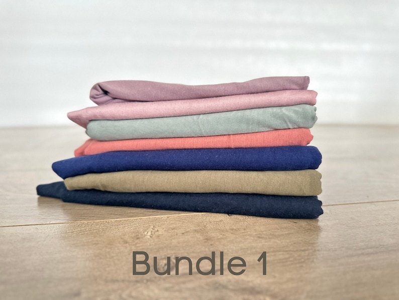 May include: A stack of six folded fabric squares in various colors, including pink, light blue, coral, navy blue, khaki, and dark blue. The fabric squares are arranged in a stack, with the pink square on top and the dark blue square on the bottom. The text "Bundle 1" is written on a wooden surface below the stack of fabric.