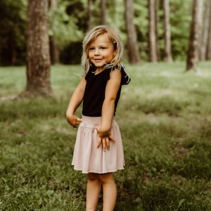 May include: A young girl with blonde hair and a black sleeveless shirt stands in a grassy area with trees in the background. She is wearing a pink skirt and is smiling at the camera.