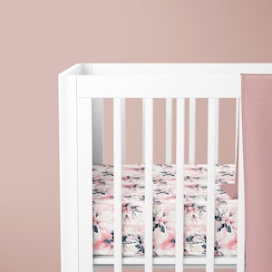 May include: A white crib with a pink floral patterned fitted sheet and a blush pink blanket draped over the side.