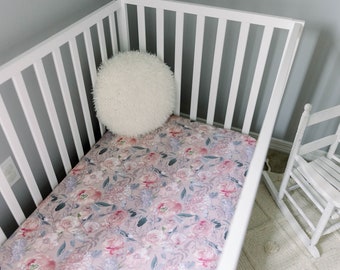 Peut inclure: Un lit bébé blanc avec un drap housse à motif floral et un oreiller blanc moelleux. Le lit bébé est dans une pièce avec une chaise berçante blanche.