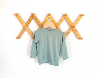 Puede incluir: Una sudadera de manga larga de color verde salvia colgada en un perchero de madera.